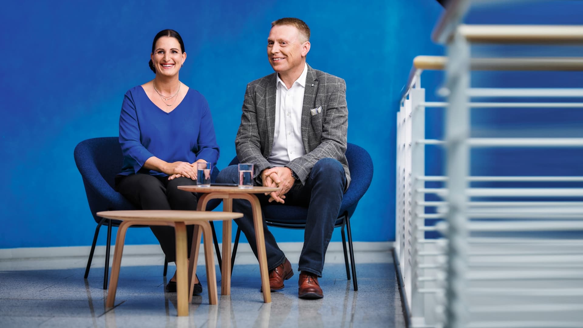 Zwei Personen sitzen vor einer blauen Wand an einem kleinen Tisch mit einem Glas Wasser und lächeln.