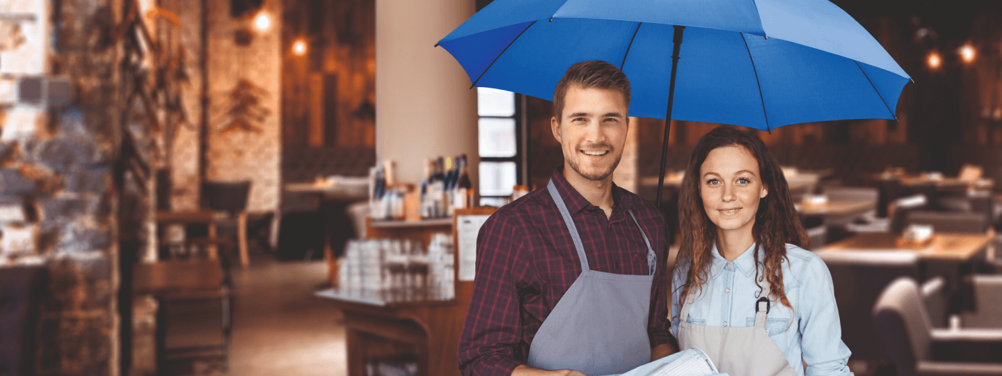 Eine junge Frau und ein junger Mann aus der Gastronomie stehen in ihrem Lokal unter dem blauen Schutzschirm.