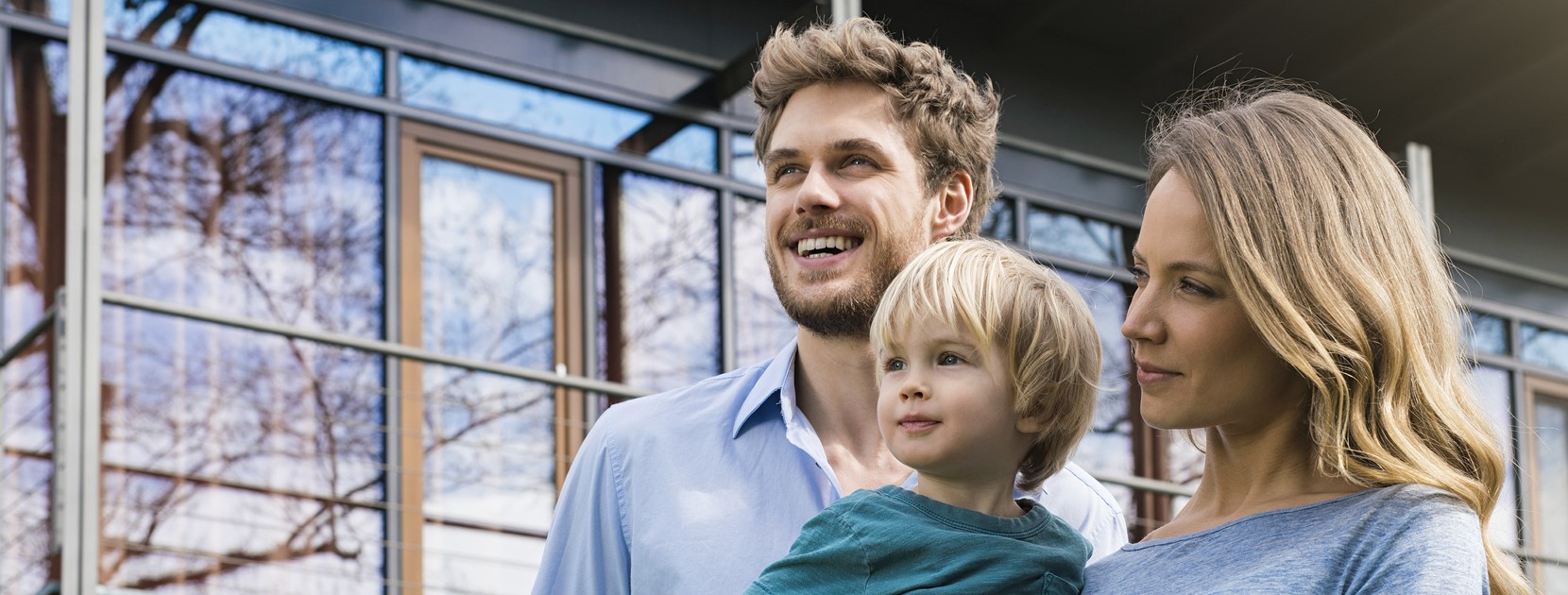 Vater und Mutter stehe vor einem Haus und haben ihren Sohn auf dem Arm.