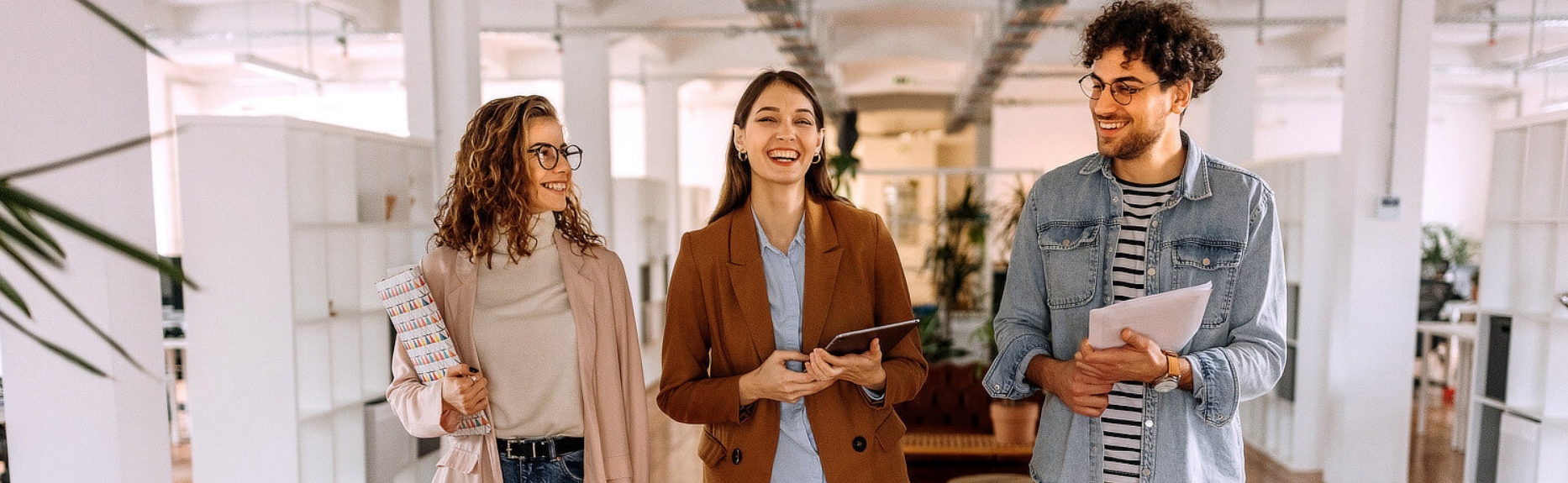 Ein junger Mann und zwei junge Frauen laufen mit Unterlagen und Tablet durch ein Großraumbüro.