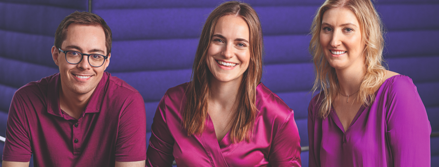 Drei Konzernmitarbeiter in lila Kleidung sitzen vor violettem Hintergrund