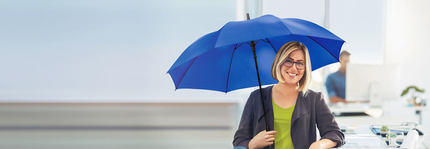Junge, lächelnde Frau mit dem blauem Schutzschirm in der Hand an ihrem Schreibtisch