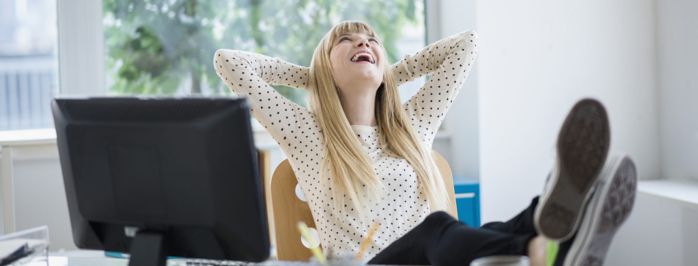 Junge Frau verschränkt die Arme hinter dem Kopf, strahlt und hat ihre Füße auf dem Schreibtisch abgelegt