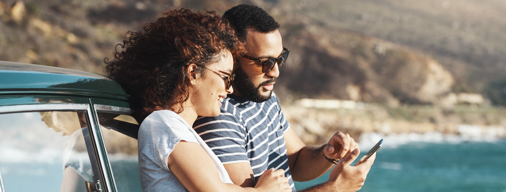 Paar schaut etwas auf dem Smartphone, am Auto gelehnt in einer Küstenregion.