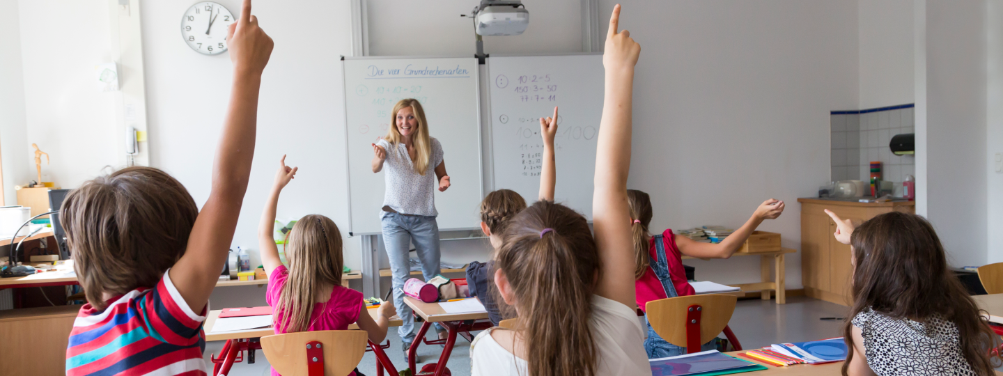 Klassenzimmer mit Lehrerin und vielen Kindern die die Hand heben