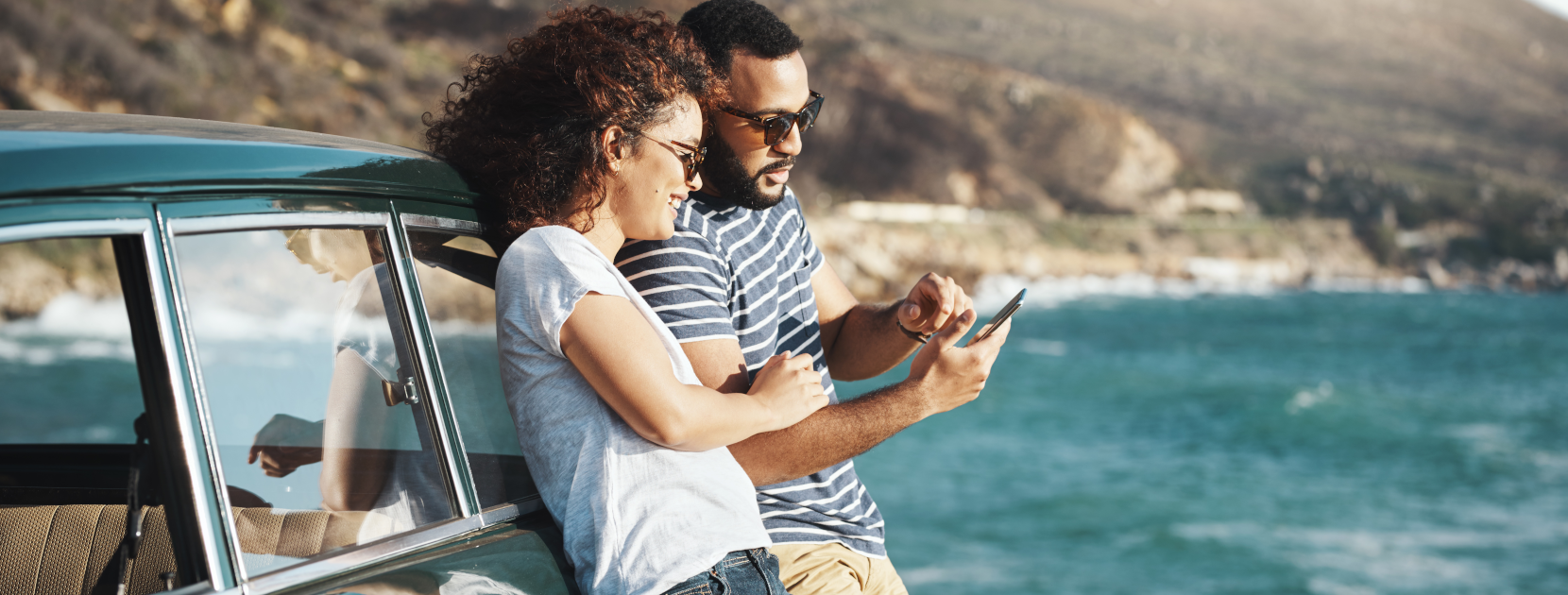 Ehepaar am Auto gelehnt schaut etwas auf dem Smartphone an einem Küstenort mit Meer.