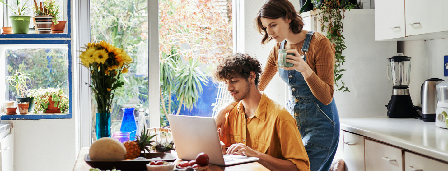 Junger Mann sitzt in der Küche am Laptop, hinter ihm steht seine Partnerin und schaut ihm über die Schulter.