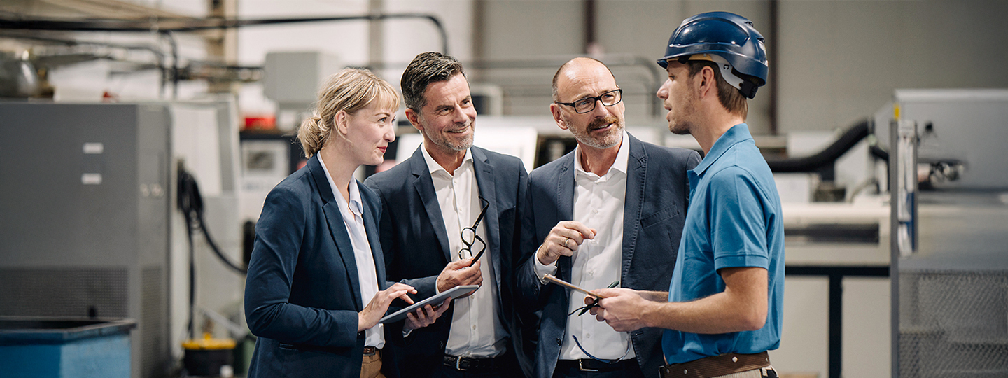 Ganz links eine Frau, daneben zwei Männer und ganz rechts ein Mann mit blauem Helm in einer Produktionsanlage
