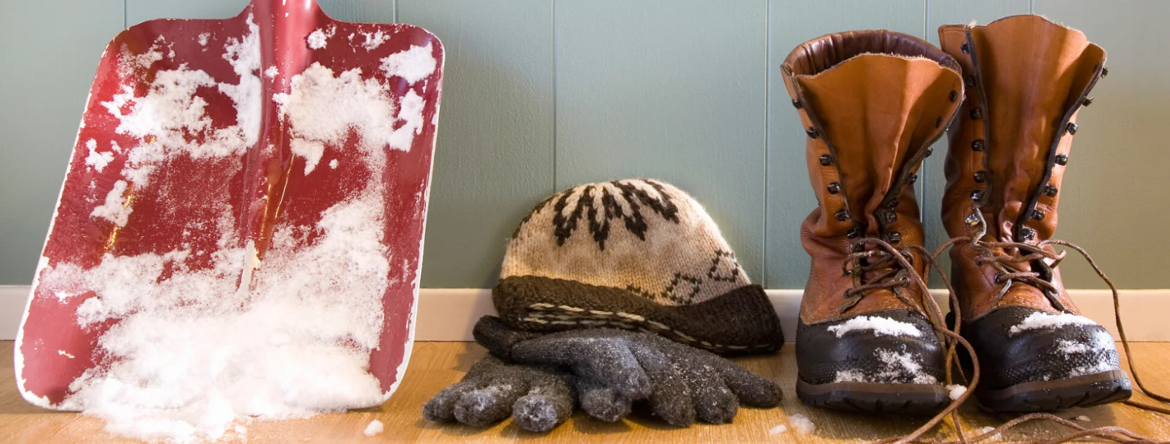 Eine Schneeschaufel mit Schnee, Mütze, Handschuhe und Schneebedeckte Stiefel stehen im Hauseingang