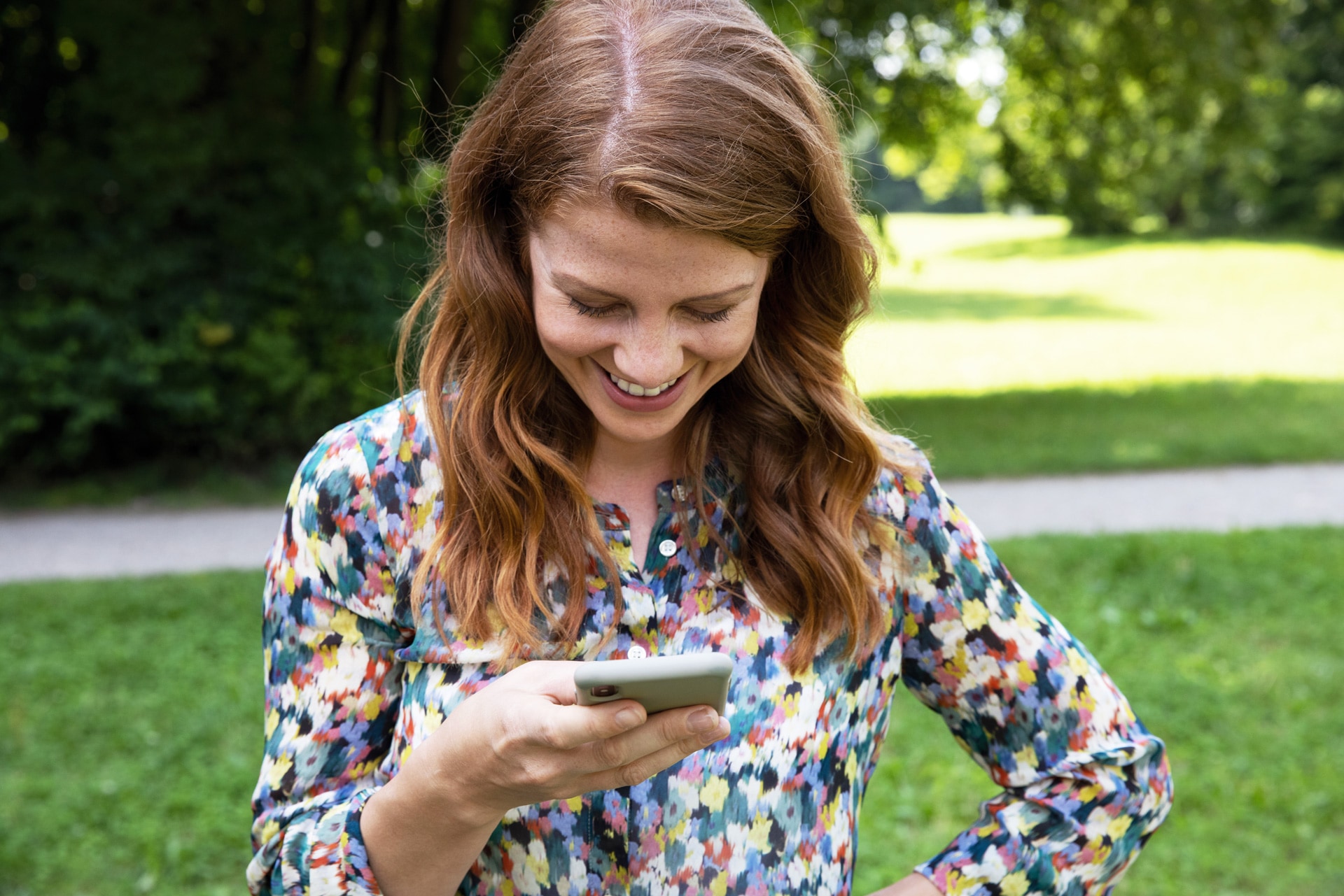 Junge Frau steht im Park und schaut grinsend auf ihr Mobiltelefon