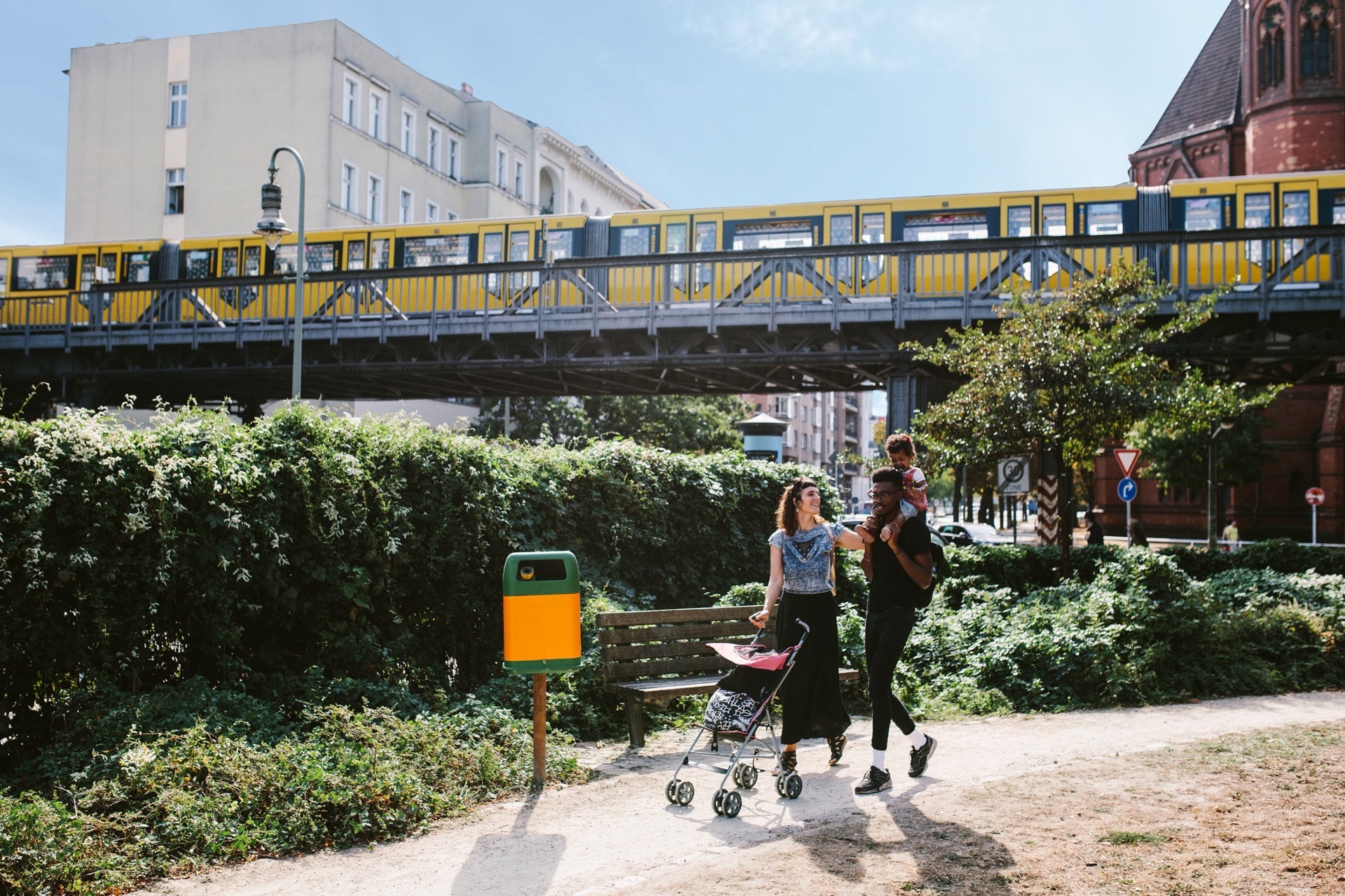 Eine junge Familie spaziert mit Kinderwagen durch einen Parkabschnitt während im Hintergrund eine S-Bahn vorbeifährt
