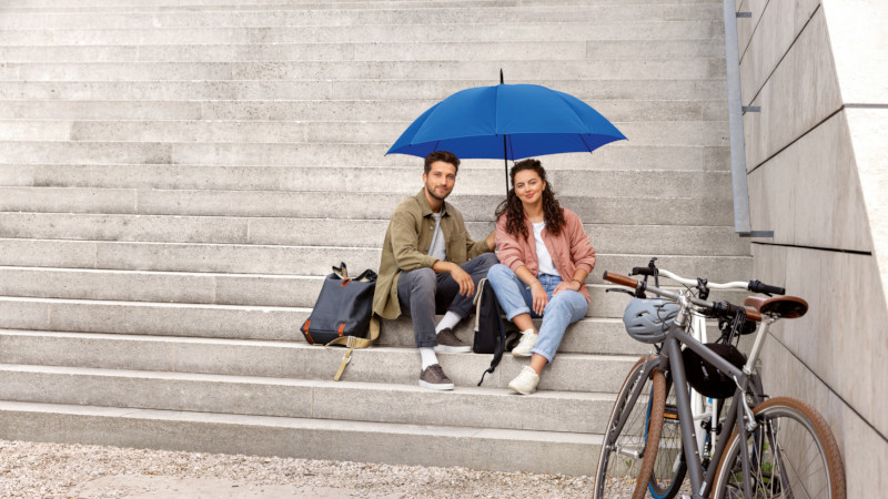 Zwei Personen auf einer Treppe sitzend mit einem blauen Schirm über ihnen und einem abgestellten Fahrrad vor ihnen